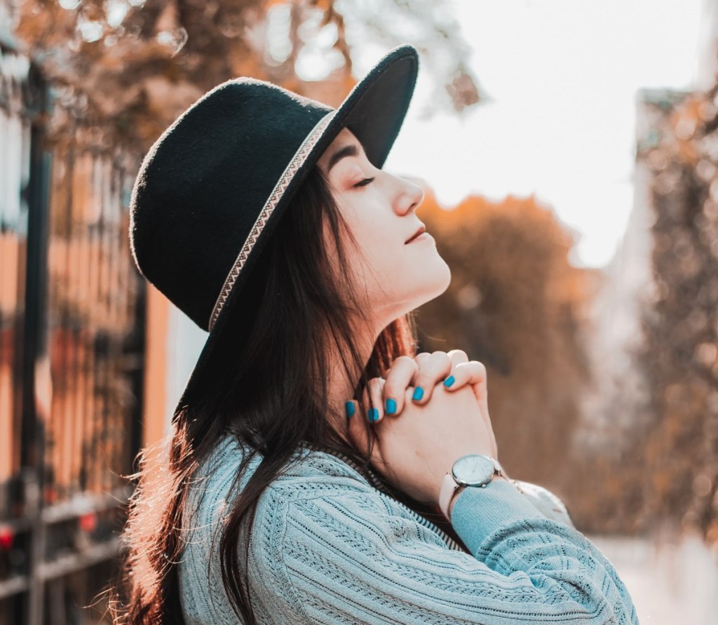 a woman in a hat and blue nail polish that matches her sweater praying with her eyes closed